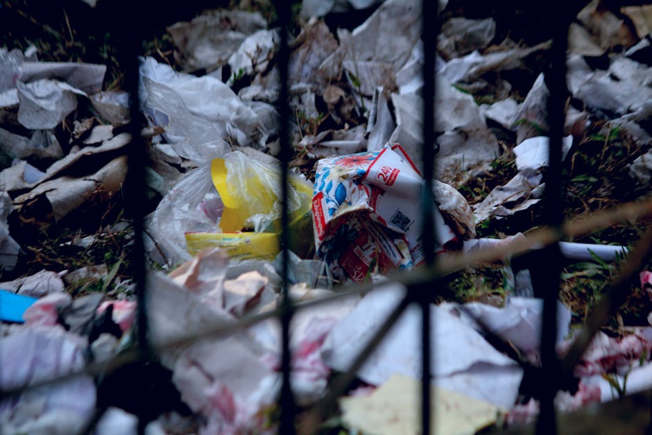 A close-up view of a pile of scattered household waste behind a black metal fence, including crumpled paper bags, plastic packaging, and food wrappers. The waste is resting on soil with sparse grass,